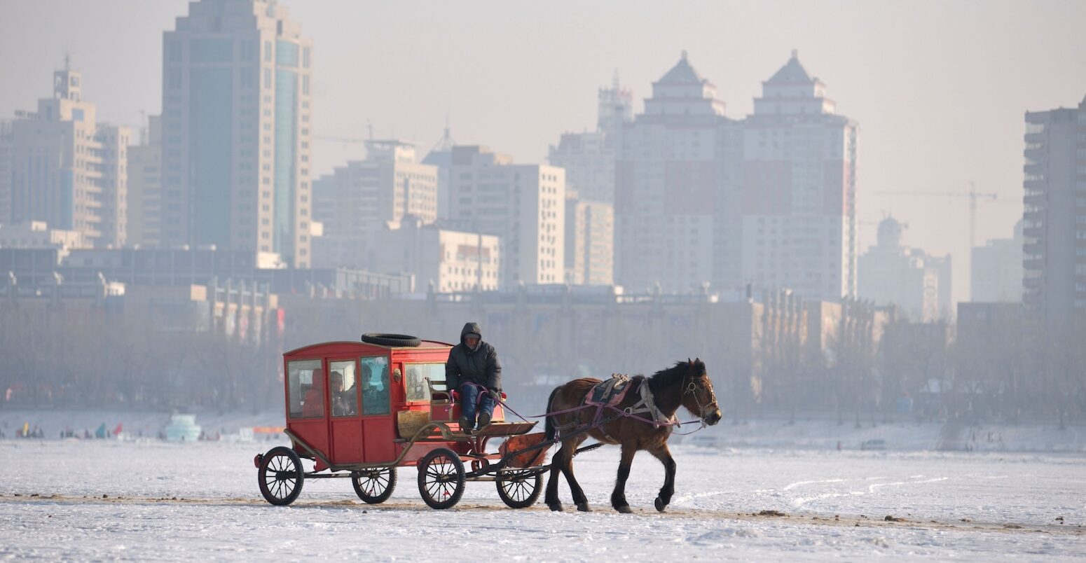 Horse and carriage crosses the frozen Songhua River with the city of Harbin in background. Heilongjiang Province, China.